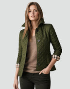 A woman models a green quilted jacket with cuffed sleeves revealing a plaid lining, paired with a brown sweater and dark green pants.